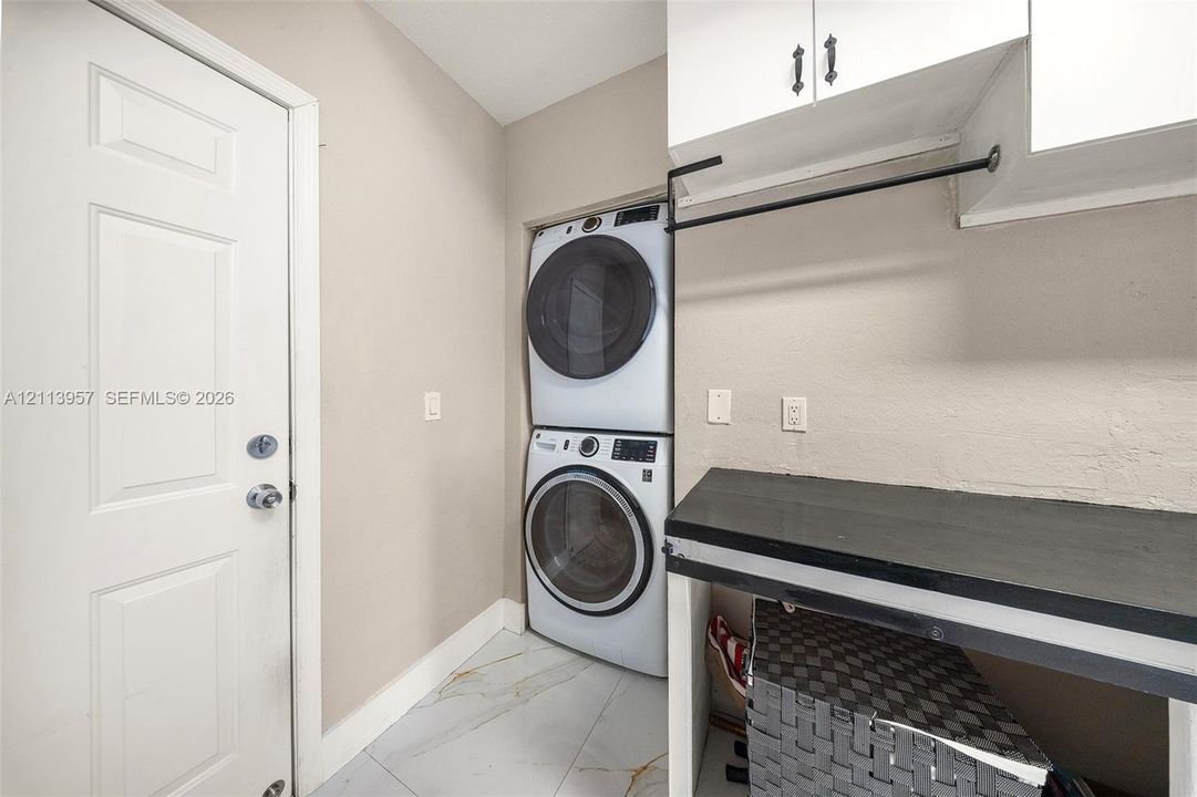 Utility area featuring large-format tile flooring, a stackable laundry unit, overhead white cabinetry, a built-in folding counter, and a wall-mounted hanging rod