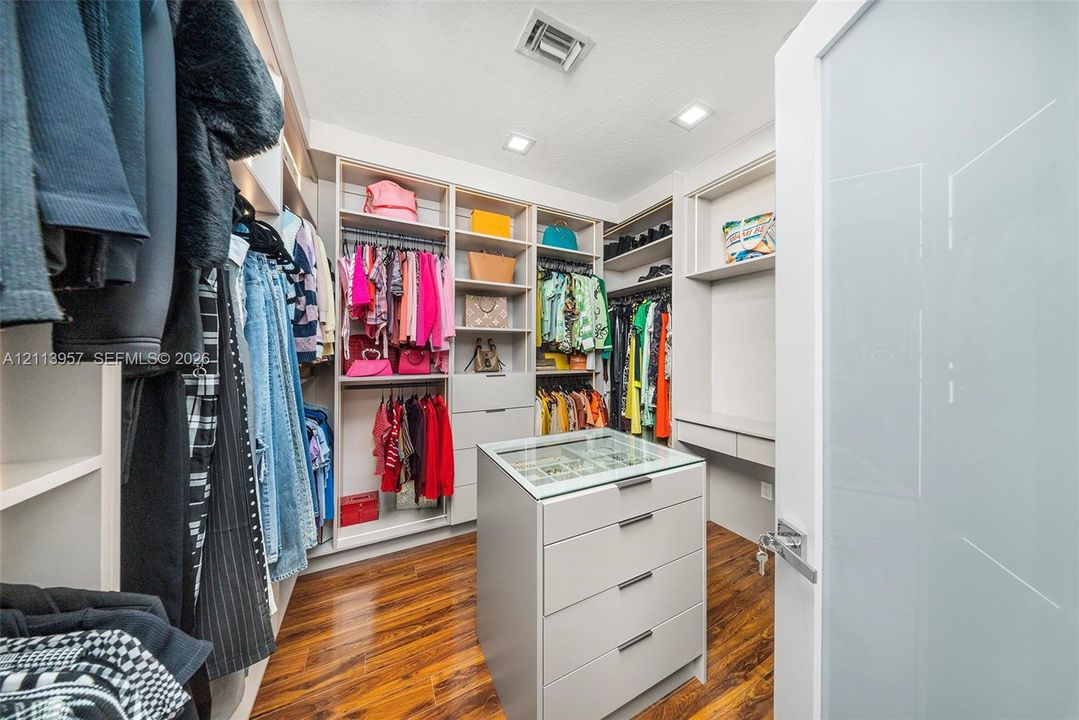 Walk-in closet featuring custom built-in cabinetry, center island with glass-top drawers, recessed lighting, and wood-finish flooring