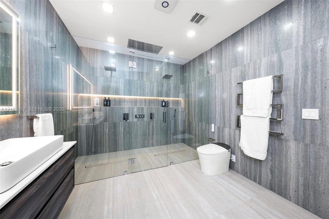 Full-height grey stone tiling, glass-enclosed walk-in shower with dual overhead fixtures and recessed lighting, wall-mounted commode, and dark-finish floating vanity with vessel sink