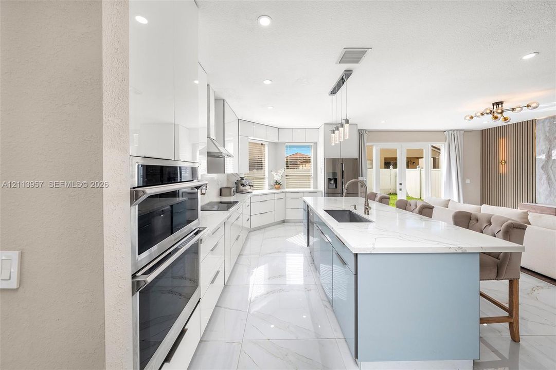 Kitchen featuring high-gloss white cabinetry, a center island with stone-surface countertop, stainless steel double wall ovens, large-format tile flooring, and integrated pendant lighting
