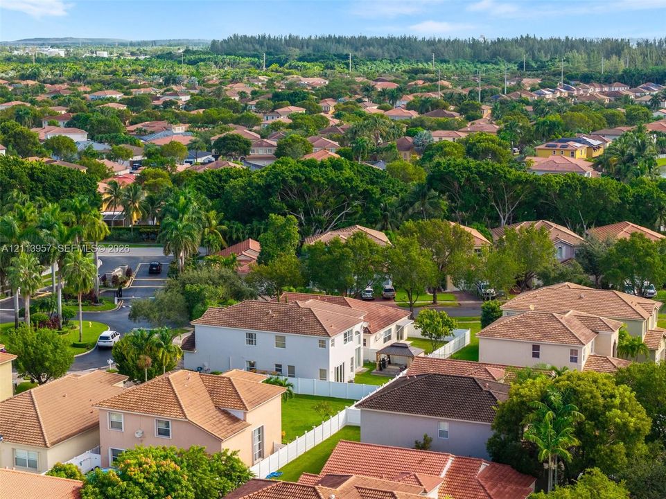 Aerial perspective of a suburban neighborhood featuring paved roadways, multi-level residential structures with tile roofing, white perimeter fencing, and mature canopy trees
