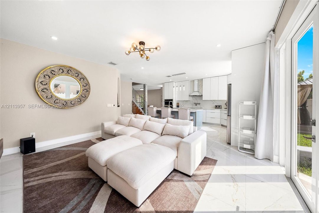 Open-concept interior featuring large-format tile flooring, modern multi-arm light fixture, kitchen with white cabinetry and waterfall-edge island, and floor-to-ceiling glass door access to outdoor space