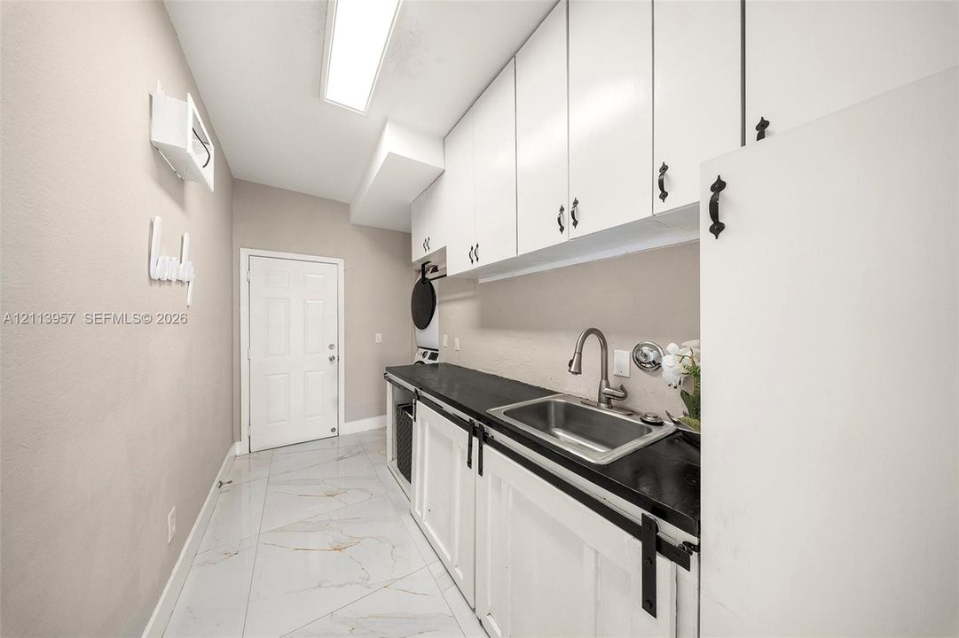 Laundry space featuring expansive white upper cabinetry, a solid-surface dark countertop, a stainless steel basin with an arched faucet, and stone-finish tile flooring