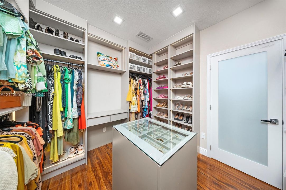 Custom walk-in closet featuring extensive built-in shelving, hanging rods, an integrated central island with a glass-top jewelry display, wood-finish flooring, and a frosted glass panel door