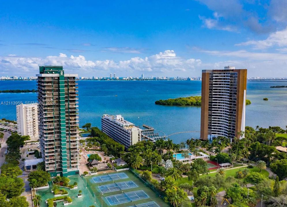 Aerial perspective of a coastal high-rise property featuring multiple tennis courts, a swimming pool, surrounding landscaped grounds, and direct water frontage