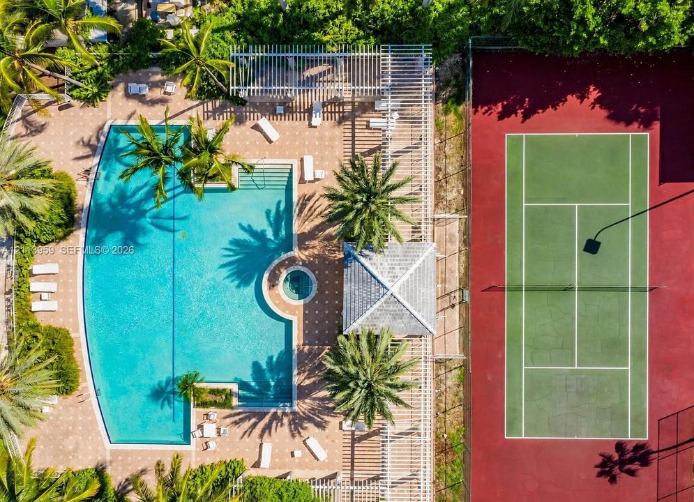 Aerial perspective featuring a swimming pool with integrated spa, a tennis court, a shaded pergola, and mature palm trees
