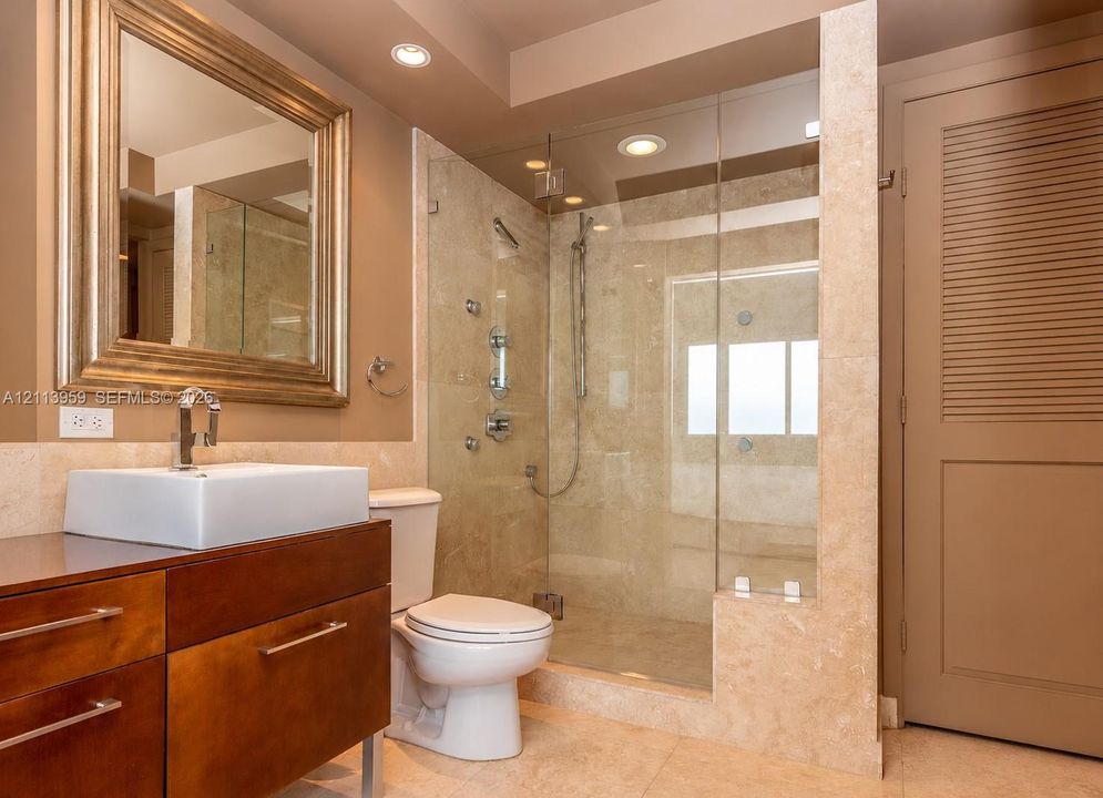 Floating wood-finish vanity with a rectangular vessel sink, frameless glass-enclosed shower with body sprays, and stone-tile surrounds