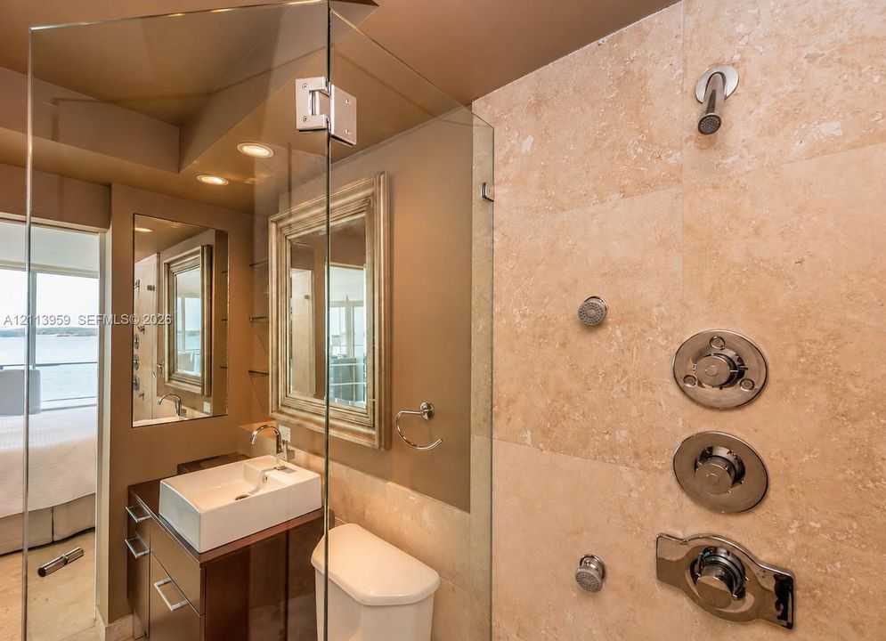 Glass-enclosed shower with stone tile surround, integrated multi-head shower system, square vessel sink with wall-mounted faucet, and custom vanity cabinetry