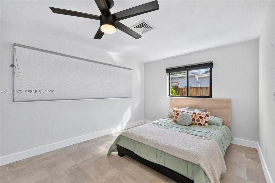 Neutral-toned tile flooring, matte black ceiling fan with integrated lighting, and dual window treatments including a roller shade and a horizontal blind