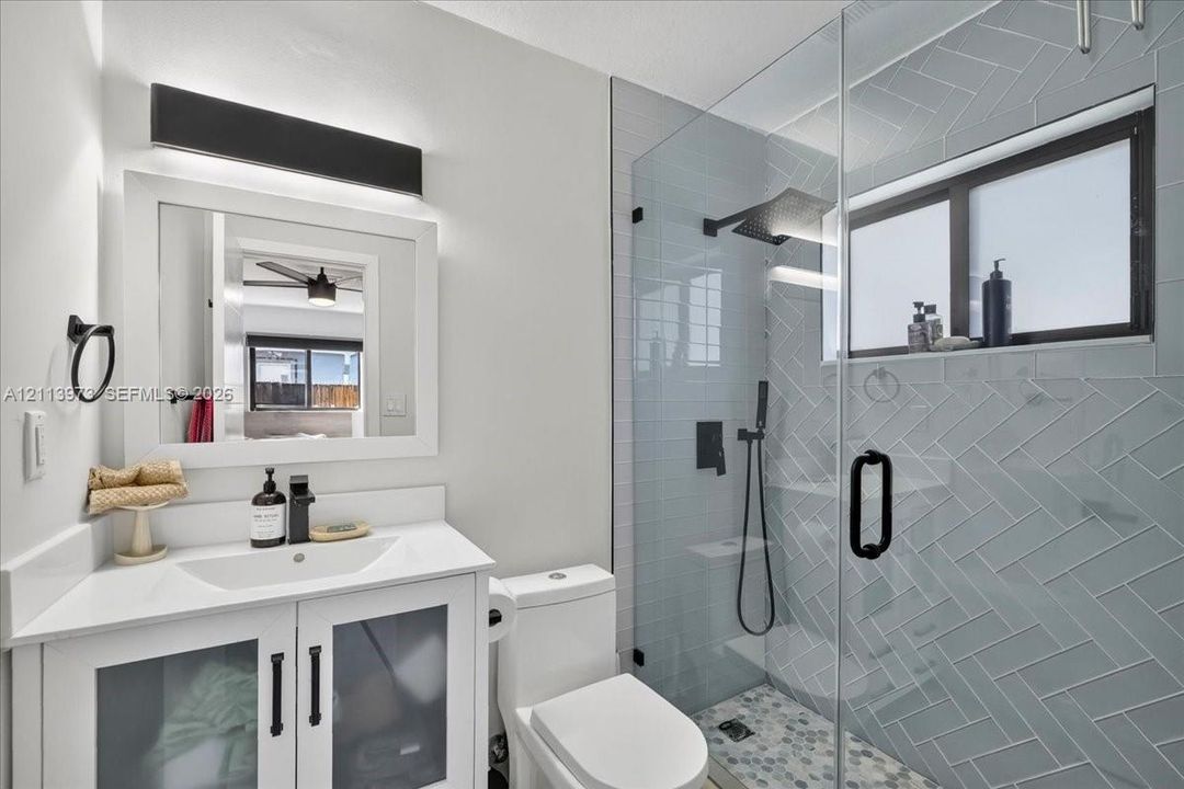 White vanity with a rectangular sink, herringbone-patterned wall tile, glass-enclosed shower with matte black fixtures, and custom light fixture