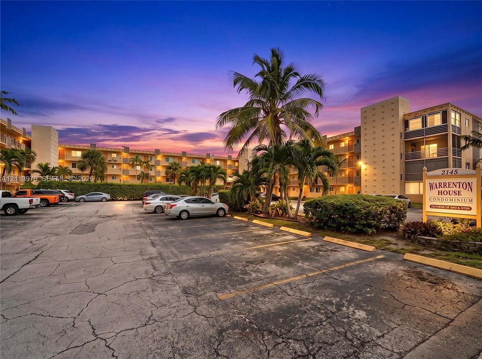 Multi-story condominium exterior featuring an on-site parking area, structured balconies, and mature palm trees