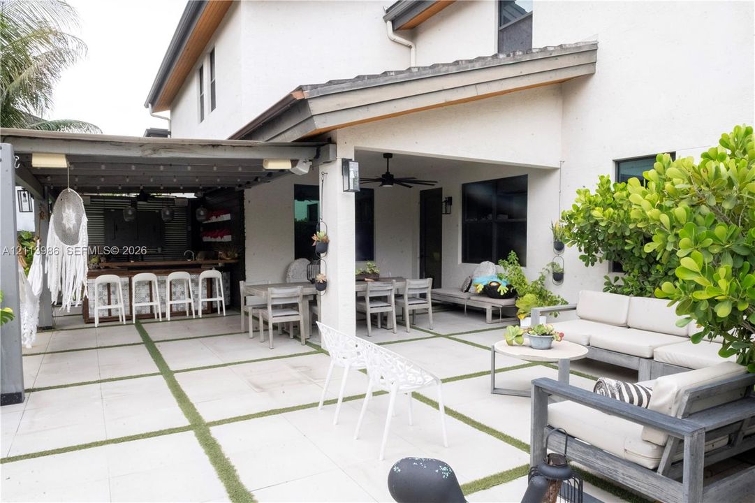 Covered patio featuring a built-in outdoor bar, large-format paver hardscaping with turf infill, a ceiling fan, and exterior wall-mounted lighting