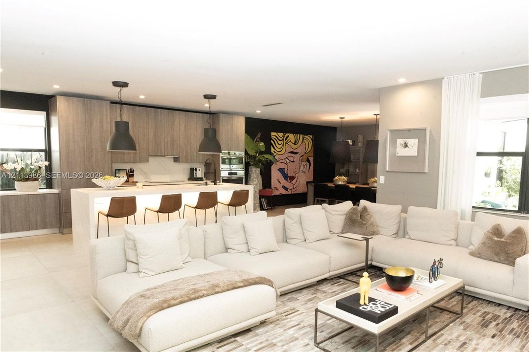 Open-concept interior featuring light wood-finish cabinetry, a large kitchen island with integrated under-counter lighting, stone slab surfacing, and matte black pendant lighting