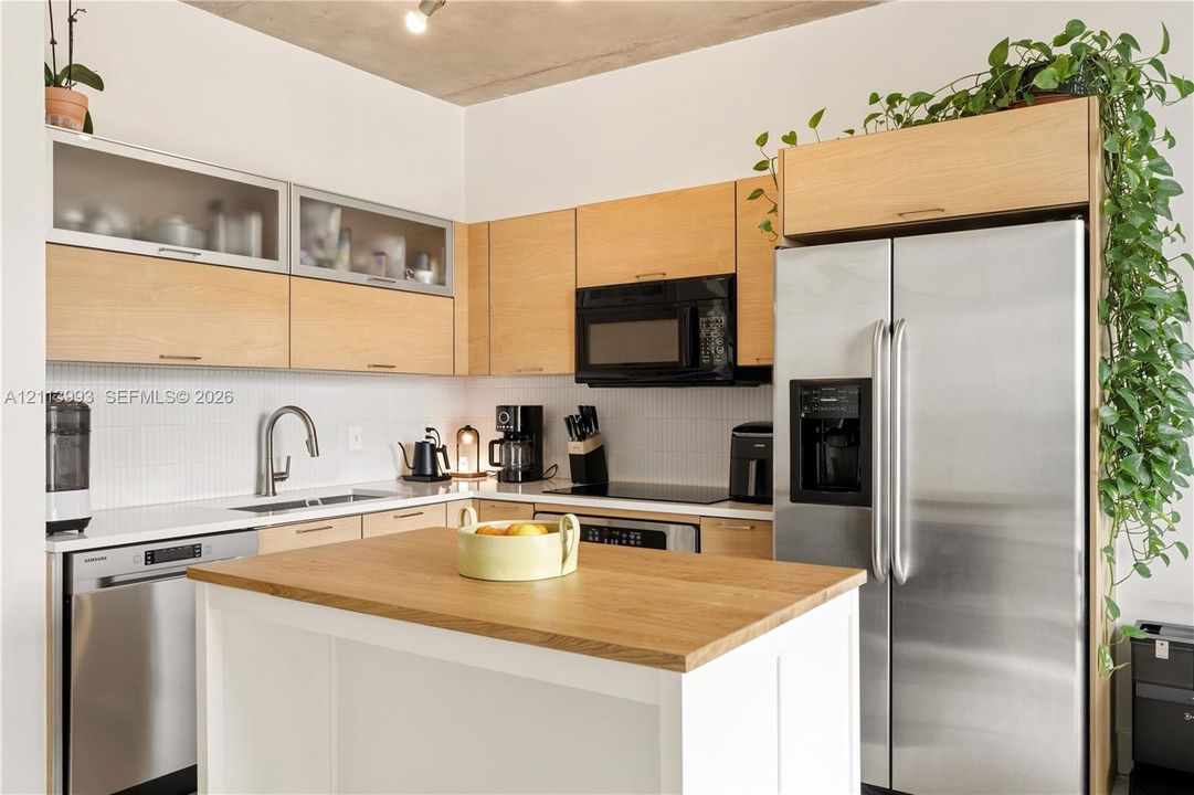 Kitchen featuring light wood-finish flat-panel cabinetry with glass-front uppers, white backsplash tile, stainless steel appliances, and a center island with a wood-finish countertop