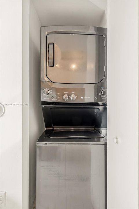 Stacked metallic laundry unit featuring a top-mounted dryer and front-loading washing machine housed within a dedicated enclosure