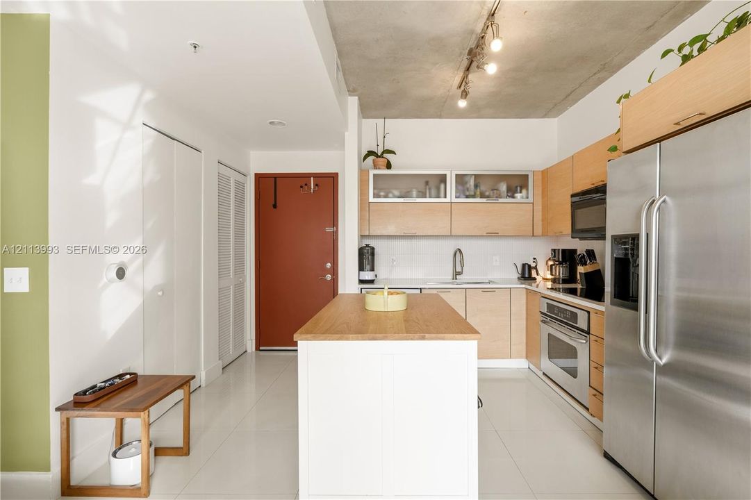 Kitchen featuring light-finish cabinetry with glass-front uppers, a central island with a wood countertop, exposed concrete ceilings with track lighting, stainless steel appliances, and tile flooring