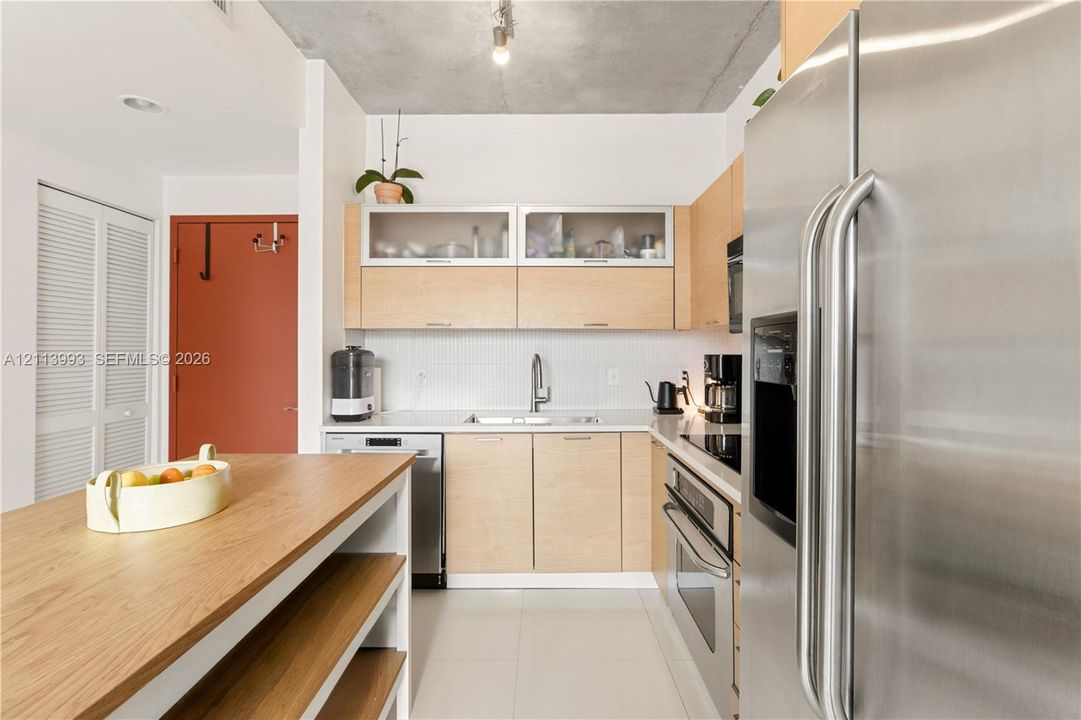 Kitchen featuring light wood-finish cabinetry, stainless steel appliances, white tile backsplash, exposed concrete ceiling, and a built-in island with open shelving