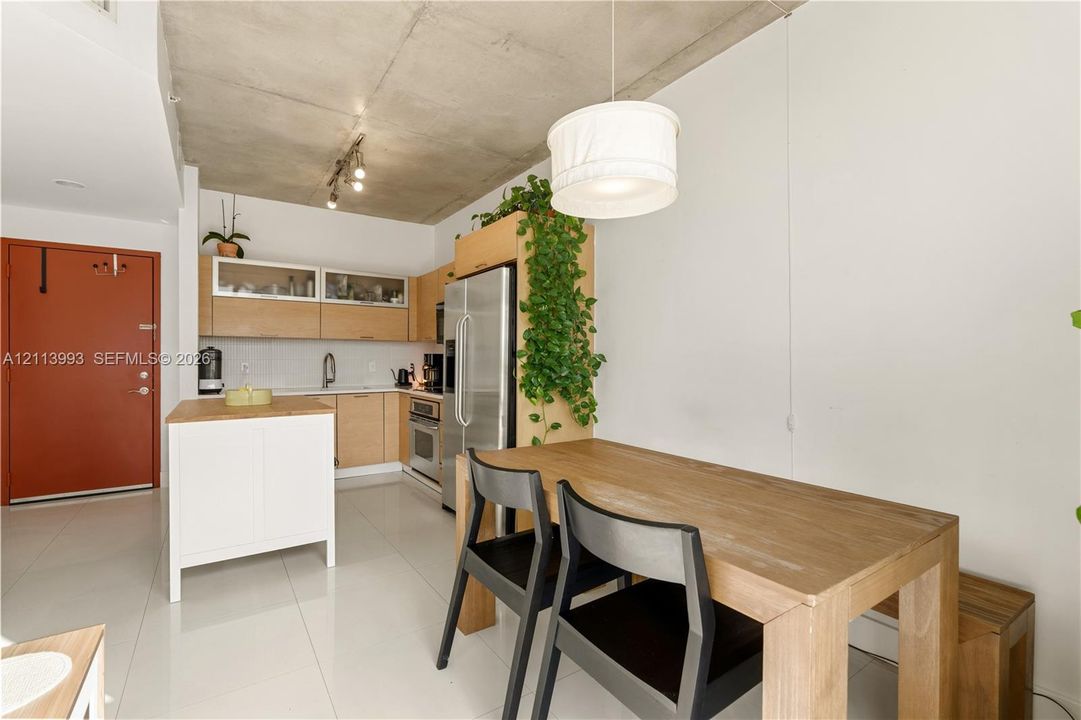 Kitchen featuring exposed concrete ceiling, light wood cabinetry with glass-front uppers, stainless steel refrigerator, white portable island, and large-format tile flooring