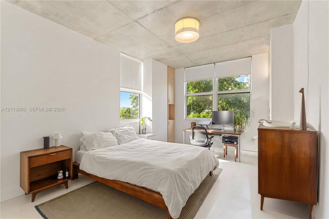 Exposed concrete ceiling, large windows with neutral roller shades, glossy white flooring, and built-in floor-to-ceiling cabinetry