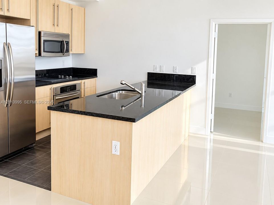 Kitchen featuring light wood-finish cabinetry, black stone countertops, stainless steel appliances, and a center island with an integrated sink and gooseneck faucet