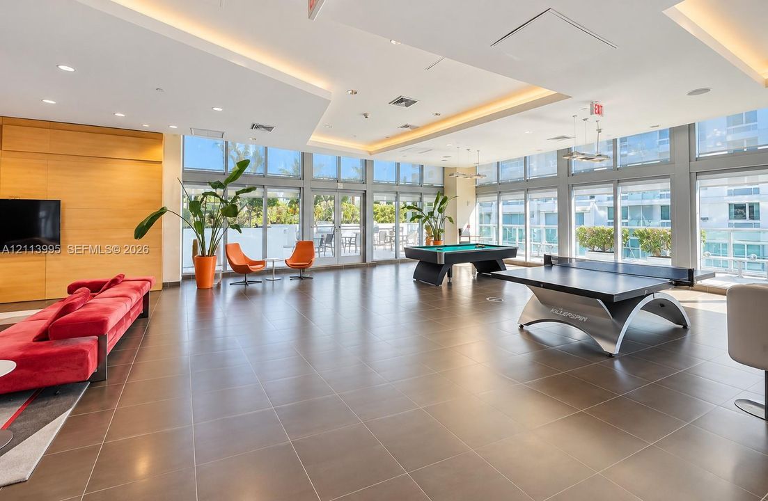 Expansive open-concept recreation room featuring large-format ceramic tile flooring, floor-to-ceiling glass windows, recessed ceiling lighting, and a wood-paneled accent wall