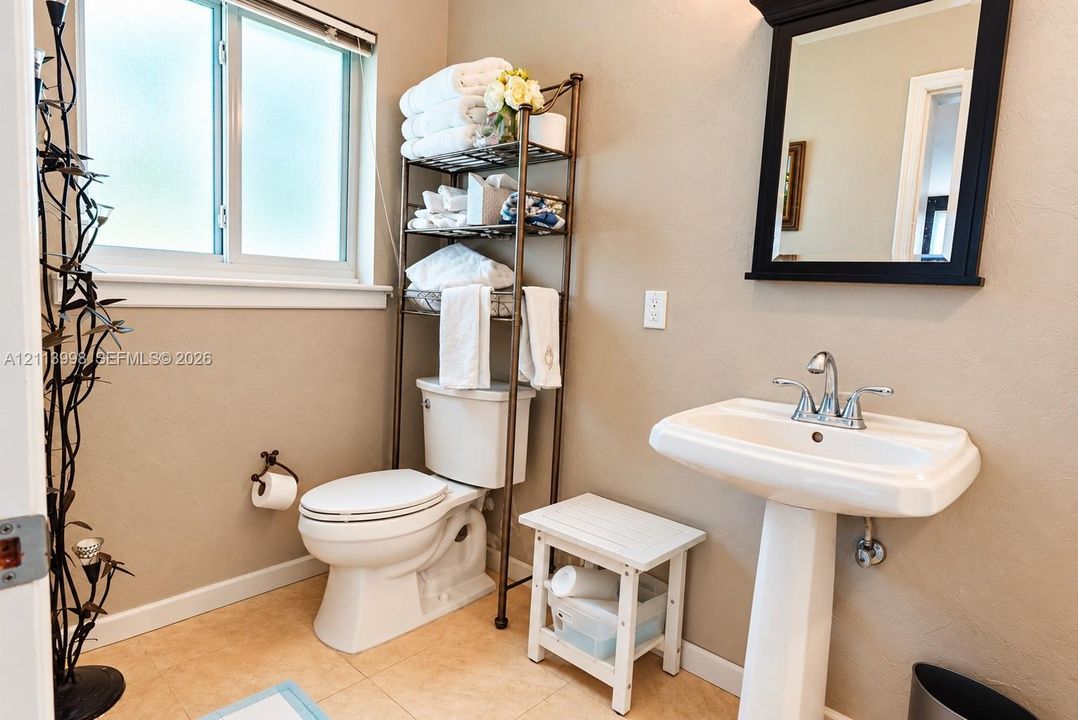 Pedestal sink with chrome dual-handle faucet, wall-mounted mirror with dark frame, privacy-glass window, and neutral tile flooring