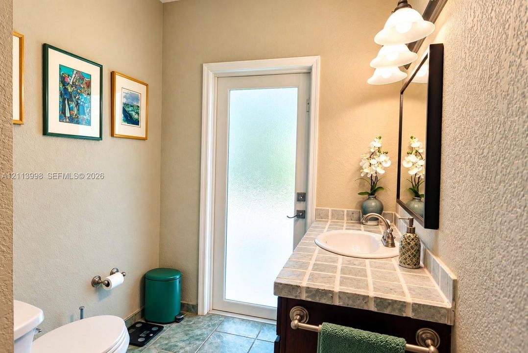 Vanity featuring a tiled countertop, undermount sink, and brushed metal faucet, complemented by a tri-light wall fixture, framed mirror, and exterior door with privacy glass