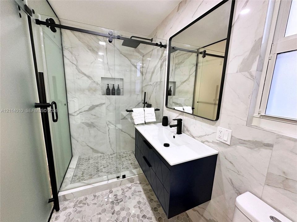 Walk-in shower featuring a sliding glass door, matte black hardware, integrated wall niche, and stone-tiled floor with a matching vanity and black-framed mirror