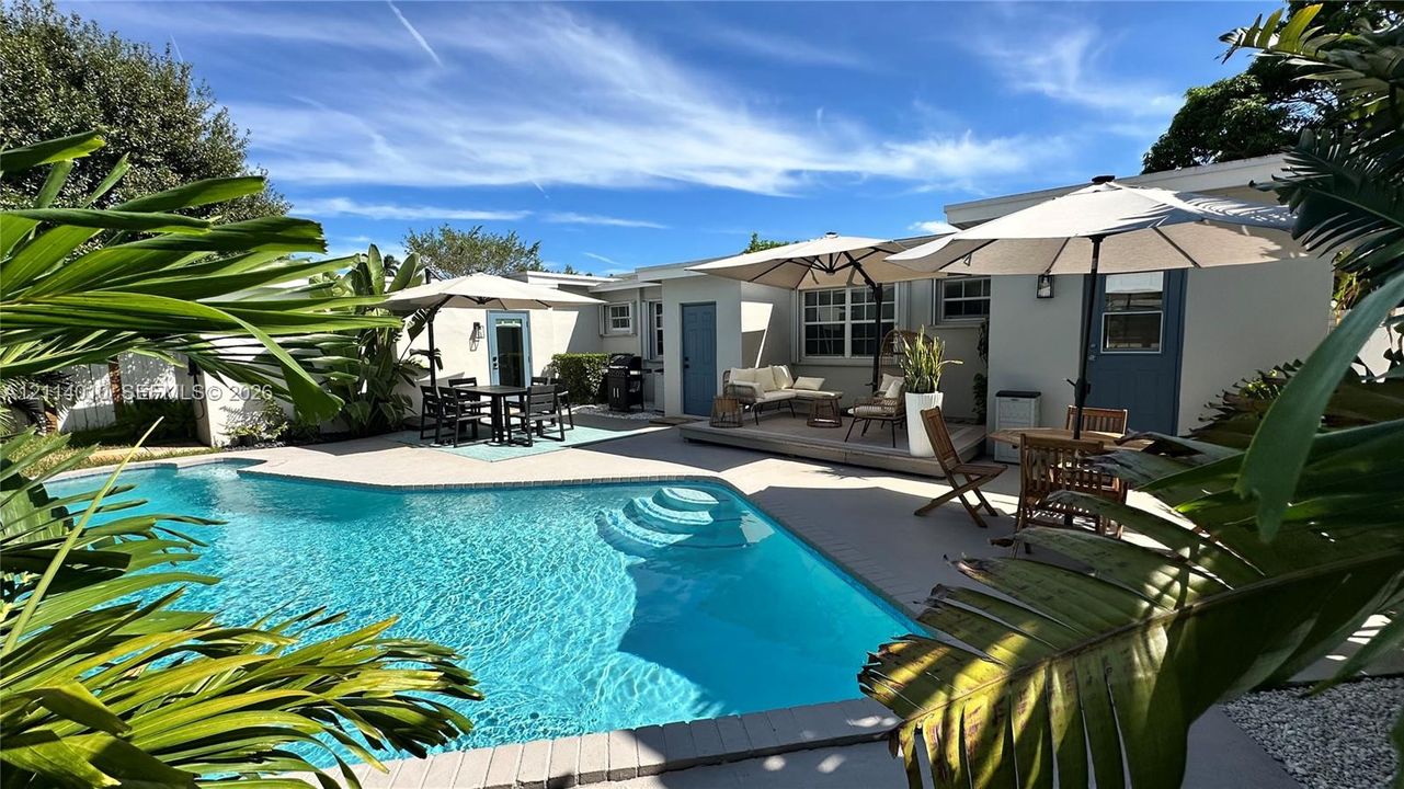 Swimming pool with integrated steps, expansive paved patio, raised wooden deck, and mature tropical landscaping