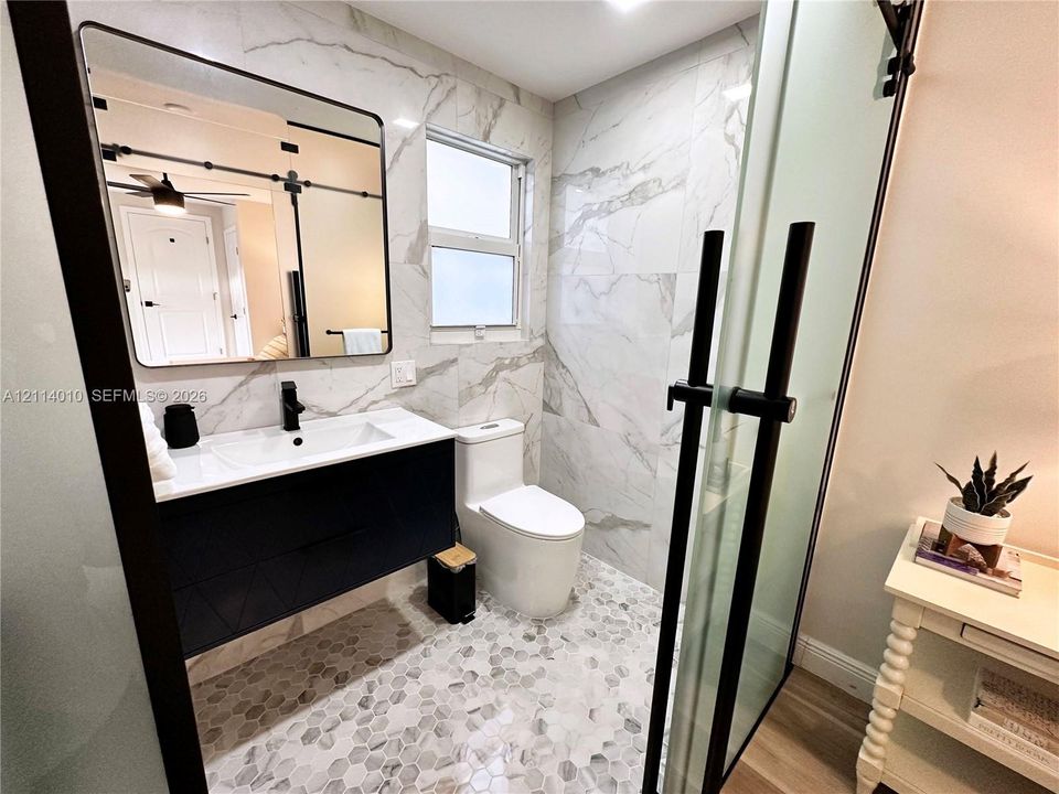 Floating vanity with a white countertop and dark cabinetry, rectangular under-mount sink with a matte black faucet, wall-mounted mirror with a black frame, full-height wall tile with stone-patterned veining, and hexagonal tile flooring