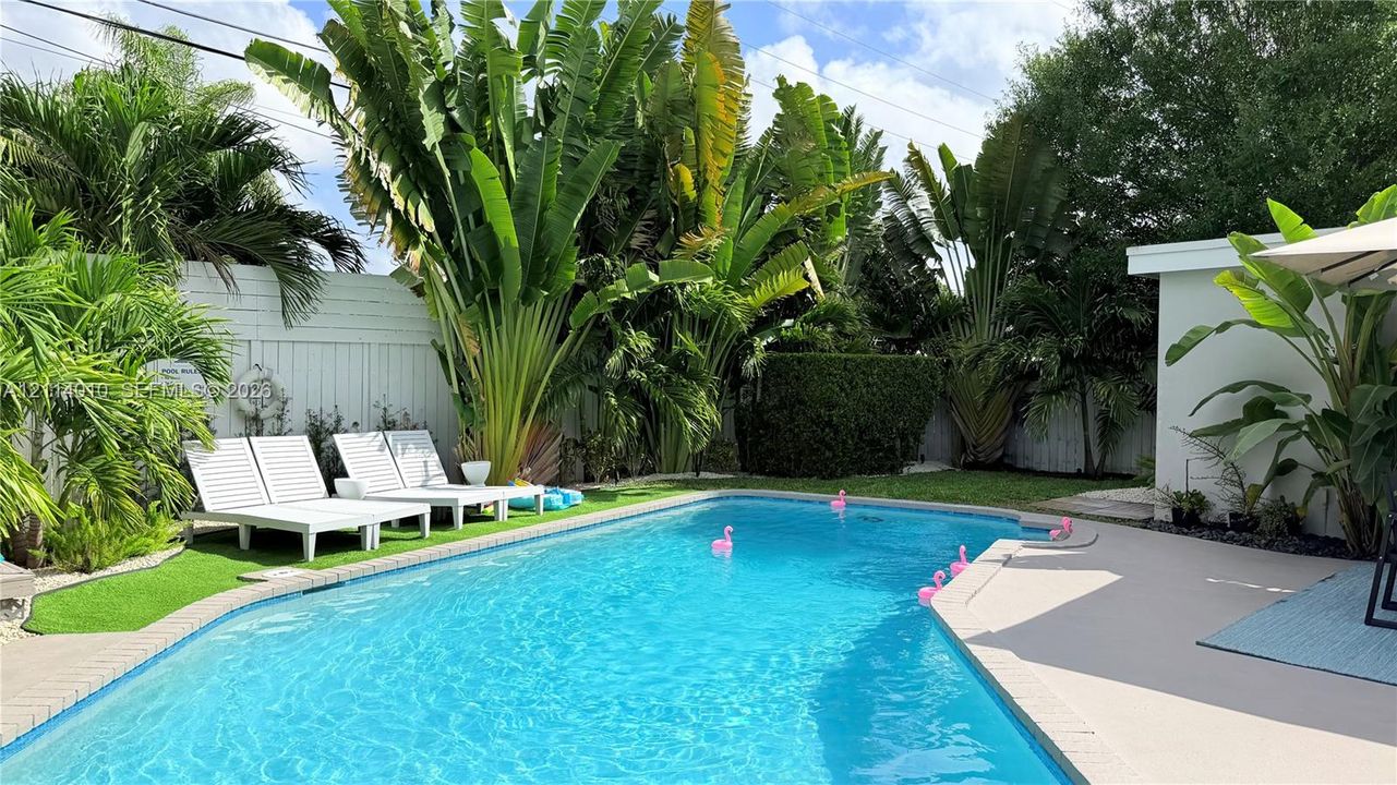 In-ground swimming pool featuring a concrete deck, enclosed by perimeter fencing, mature tropical landscaping, and artificial turf surfacing