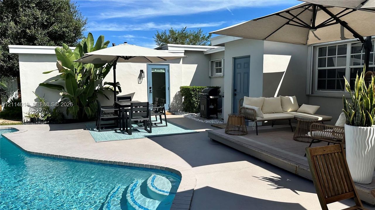 Private courtyard featuring a swimming pool with integrated steps, concrete patio surfacing, raised wood-finish deck, and light-colored exterior walls with blue-trimmed doors