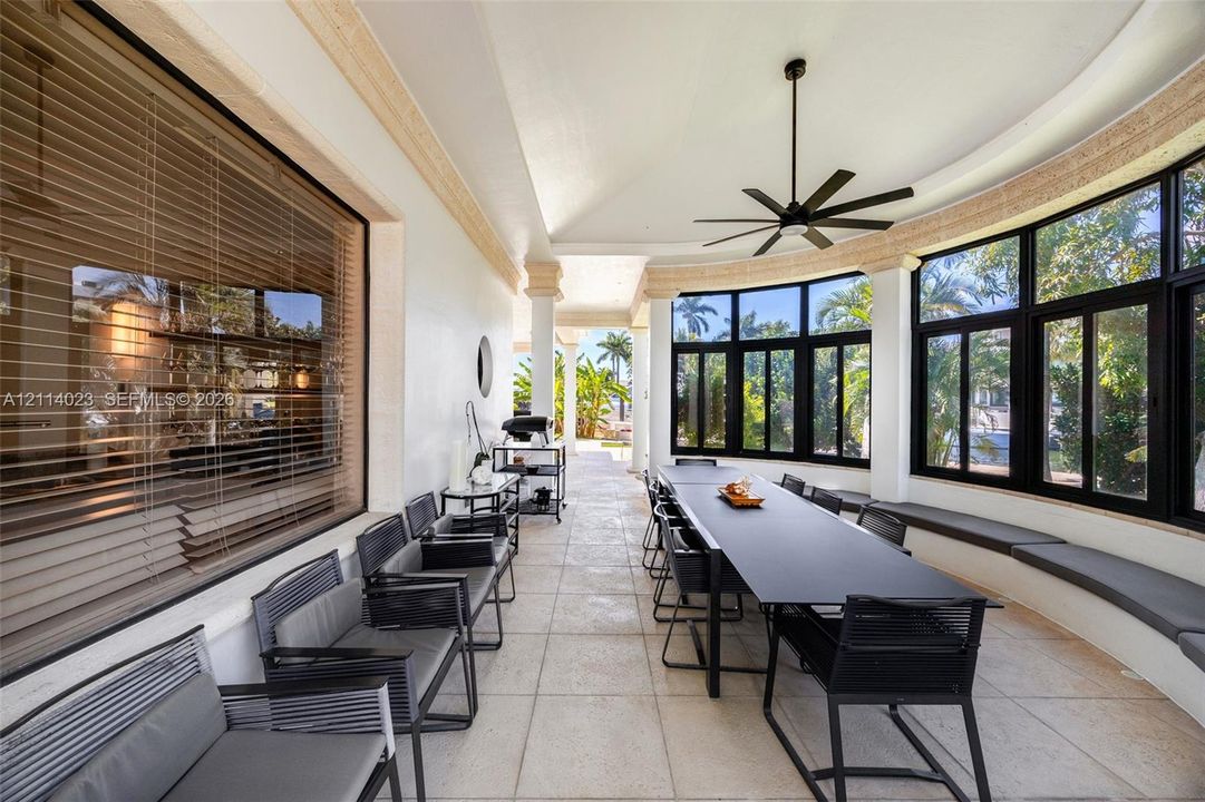Covered outdoor patio featuring light-toned tile flooring, curved architecture, floor-to-ceiling windows with black frames, built-in bench seating, and a ceiling fan