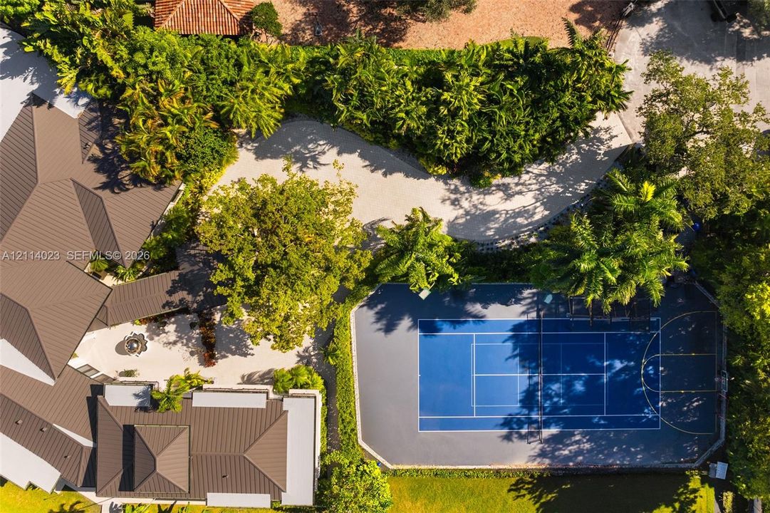 Aerial perspective of the estate featuring a multi-sport court with blue hard surfacing, expansive stone-paved driveway, metal roofing, and mature landscape canopy