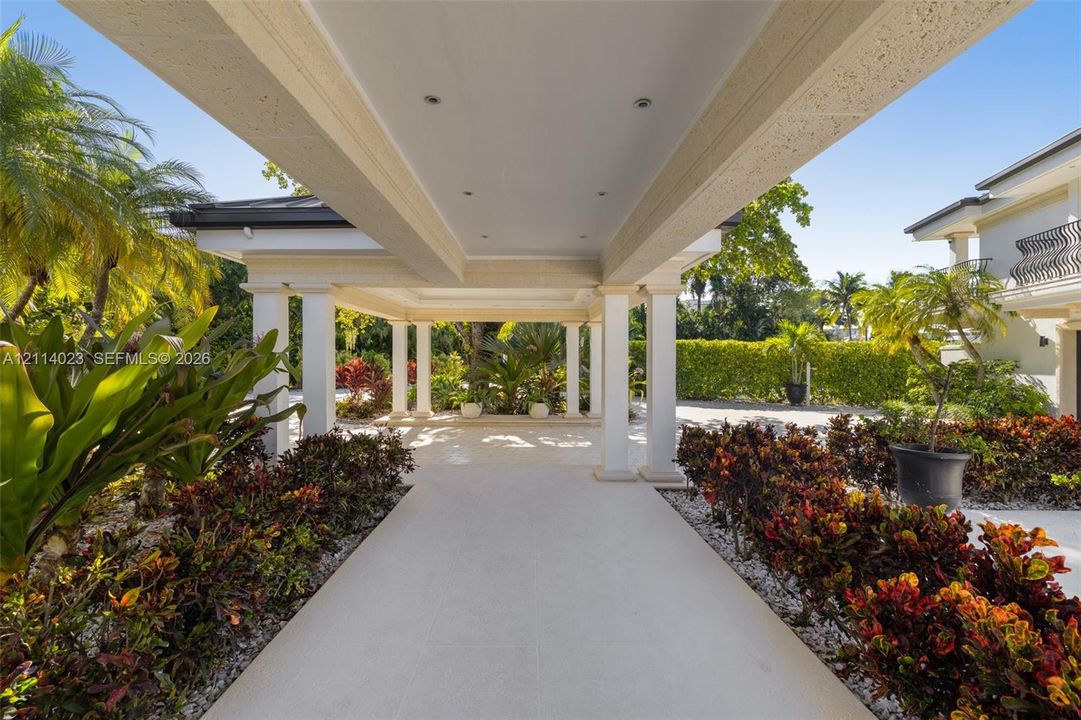Covered porte-cochère featuring a light-toned stone tile surface, structural white columns, recessed lighting, and surrounding professional landscaping