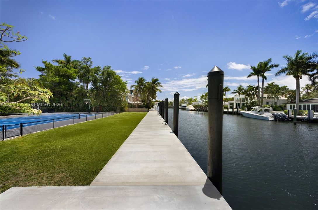 Concrete dock with mooring pilings, adjacent to a manicured lawn and a fenced sports court