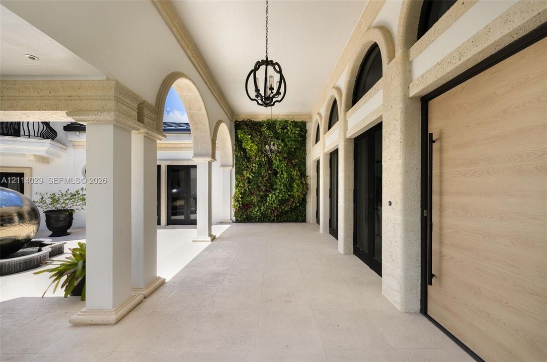 Covered loggia featuring arched architectural columns, stone-textured detailing, a large-format light-toned tile floor, a decorative hanging pendant fixture, and a vertical green garden wall