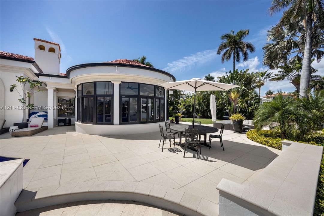 Expansive stone patio featuring a curved architectural structure, multi-pane floor-to-ceiling windows, integrated masonry seating, and a terracotta tile roof