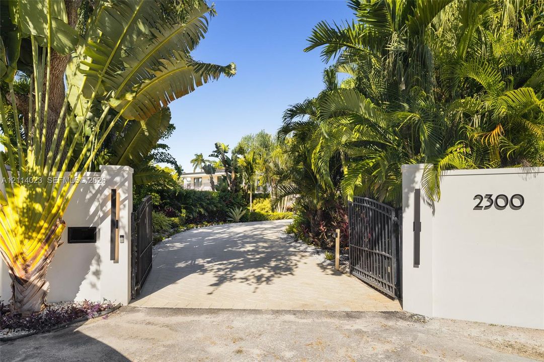 Gated entrance featuring decorative stucco walls, iron swing gates, paver driveway, and mature tropical landscaping