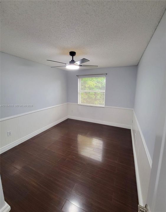 Room featuring wood-finish flooring, white wainscoting, a single-hung window, and an integrated ceiling fan with light fixture
