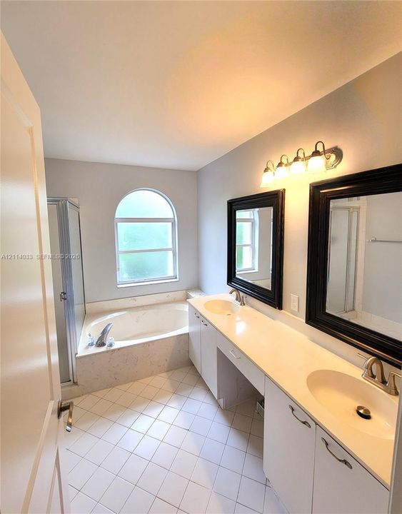 Bathroom featuring a dual-sink vanity with a solid-surface countertop, an arched window above a soaking tub with tile surround, a separate glass-enclosed shower, and tile flooring
