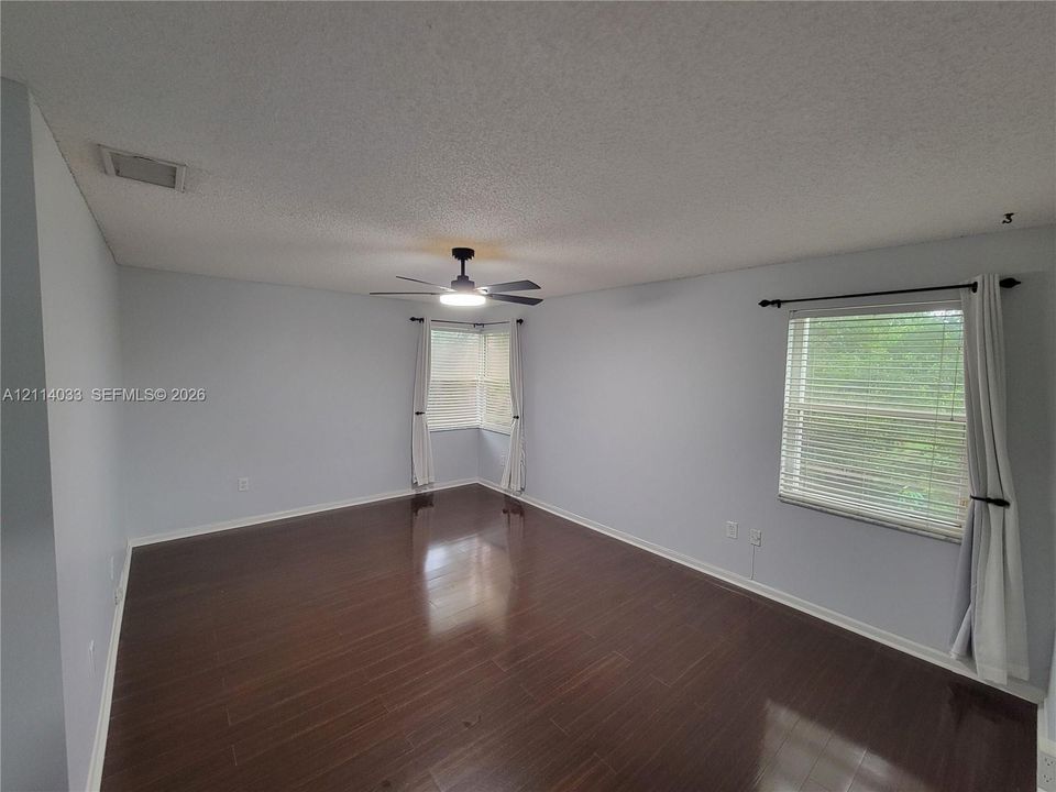 Spacious room featuring dark wood-finish flooring, a central ceiling fan with an integrated light fixture, dual-window corner configuration, and horizontal window blinds