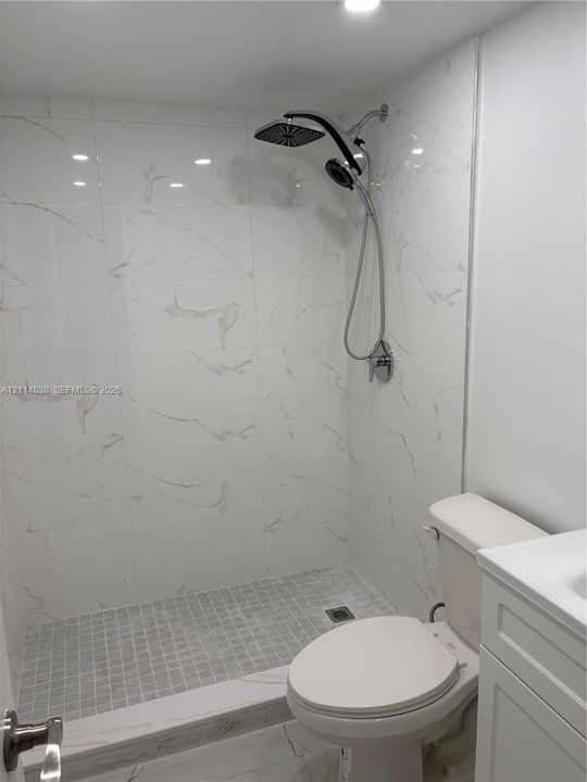 Walk-in shower featuring large-format white tile with grey veining, a mosaic-tiled shower floor, a dual-head shower fixture with a handheld attachment, and a white vanity cabinet
