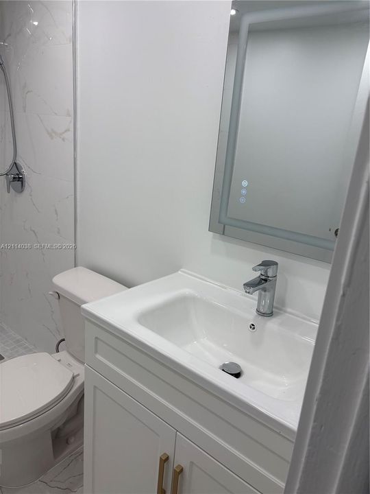 White vanity with brushed gold hardware, integrated porcelain sink, polished chrome faucet, illuminated wall-mounted mirror, and tile shower surround