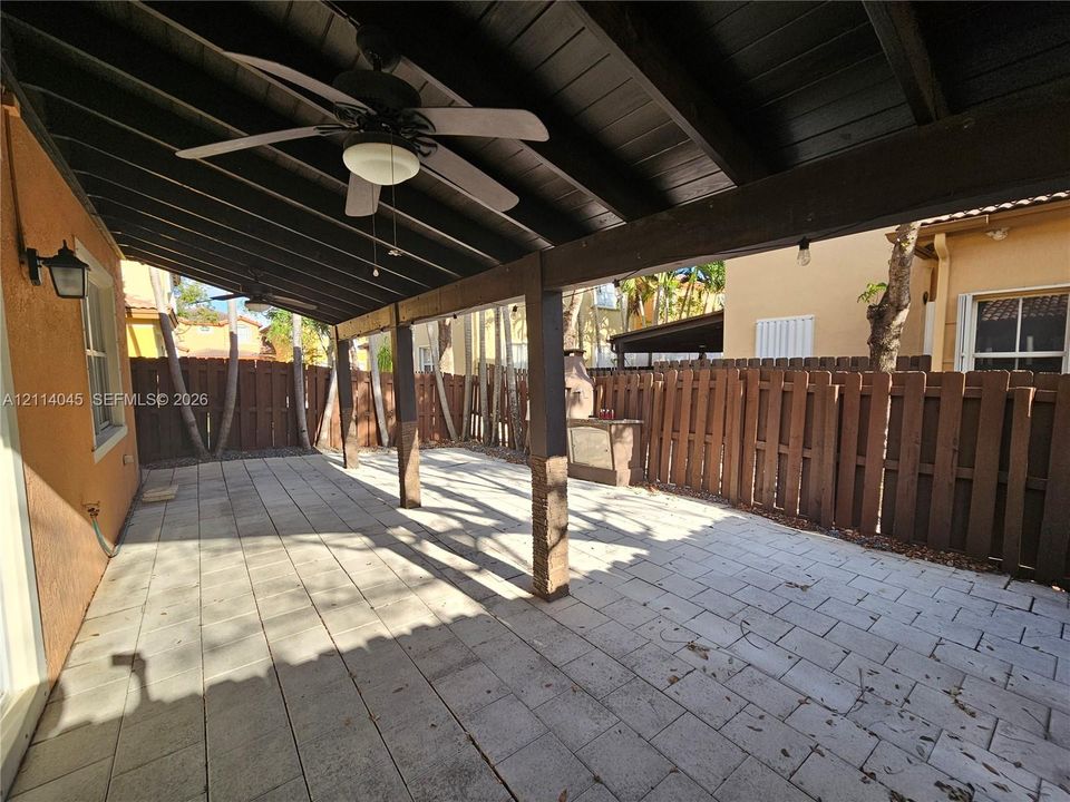 Covered patio featuring concrete paver flooring, dark-stained wood support beams, built-in ceiling fans, an exterior wall-mounted light fixture, and a vertical wood privacy fence