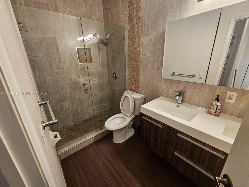 Walk-in shower with a glass enclosure, wall-mounted showerhead, and a recessed tiled niche, featuring a wood-finish floating vanity with a dual-basin countertop and integrated cabinetry