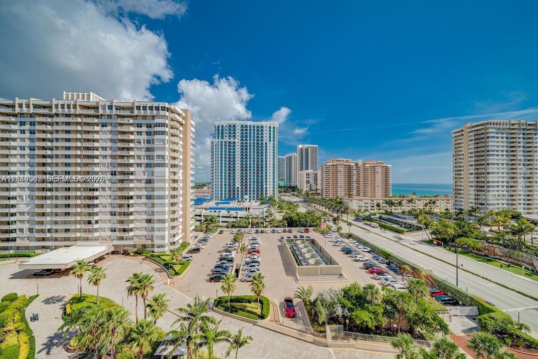 High-rise residential exterior featuring expansive surface parking, paved driveways, manicured tropical landscaping, and structural balconies