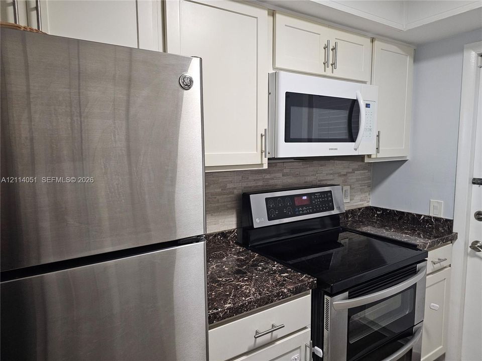 Kitchen featuring white shaker-style cabinetry, stainless steel refrigerator, dark stone-patterned countertops, stacked-tile backsplash, and integrated microwave
