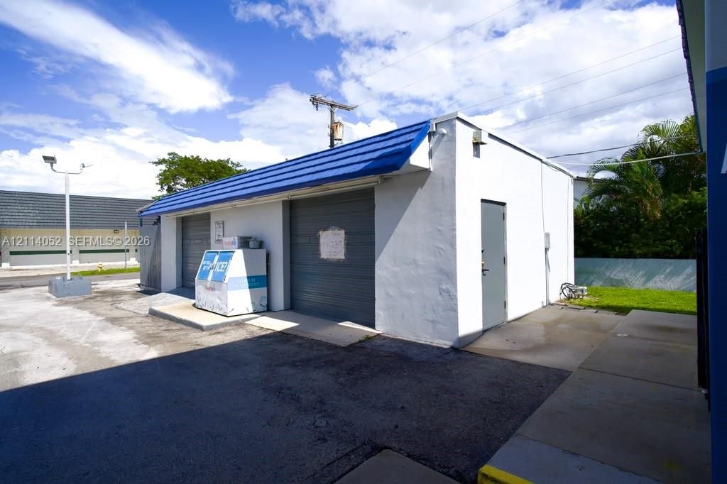 Concrete structure with two rolling garage doors, blue metal awning, exterior lighting, and asphalt paving
