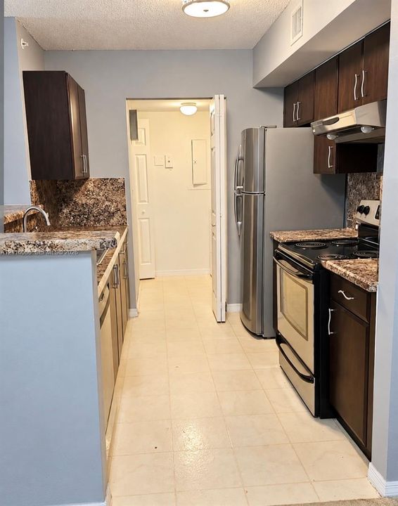 Kitchen featuring dark wood-finish cabinetry, stone-patterned countertops, stainless steel refrigerator, electric range with matching overhead hood, and tile flooring